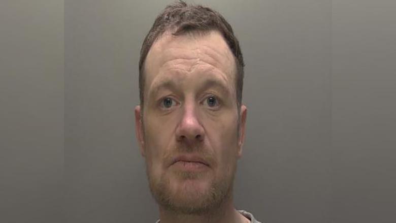 Police custody photograph of Timothy Tinson. He has short brown hair and beard and is wearing a grey T-shirt