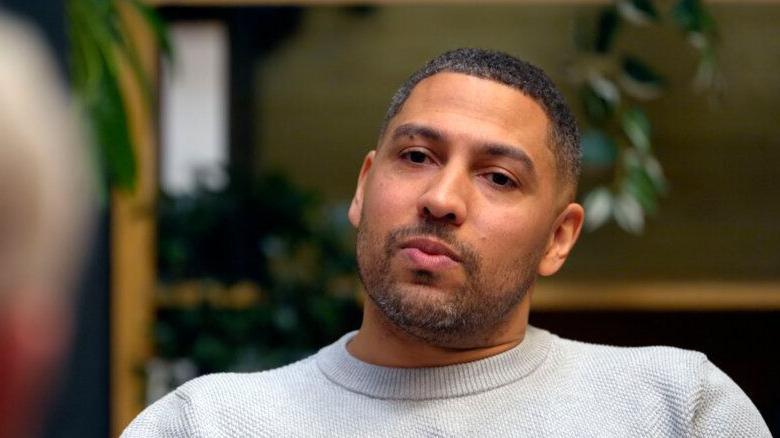 James in a therapy session, wearing a white knitted top, against a blurred interior background