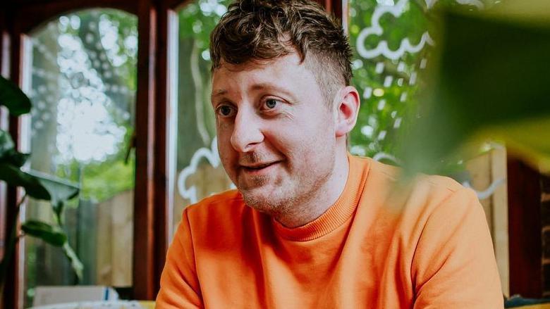 Children's author Jarvis who has brown hair wearing an orange t-shirt sitting on a sofa in front of windows with a garden view.