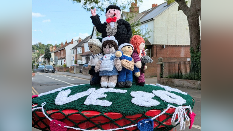 A postbox topper featuring knitted woolen figures, including Sir Winston Churchill, a nurse, a sailor and a postwoman, on the postbox, with houses visible behind it.