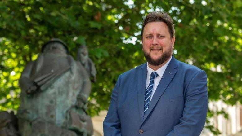 Lee Walker smiles at the camera. He has short brown hair and a dark thick beard. He wears a blue suit with a white shirt underneath and a blue tie.