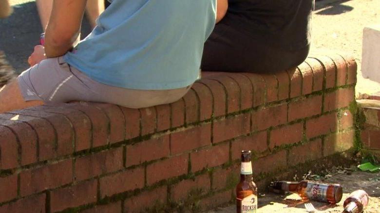 Two males sit on a small red brick wall. A number of beer bottles can be sen lying at the bottom of the wall, which is very small. The image shows both men facing away from the camera and their heads or legs can not be seen. One is wearing a green short and grey shorts while the other a black t shirt