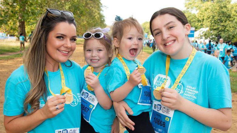 Louise Hubbard with her children Florence, Fleur and Felicity. They are all standing in a park wearing blue T-shirts, holding medals and smiling at the camera.