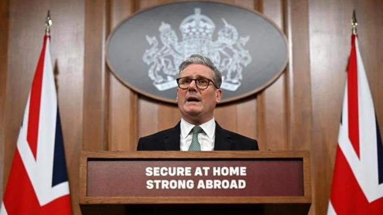 Sir Keir Starmer speaking at a podium flanked by two Union Flags, behind a podium bearing a sign which reads "Secure at home, strong abroad"