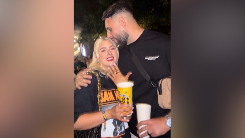 A young woman with blonde hair stands holding up her hand with several rings on it. A man with a beard stands with his arm around her kissing her on the forehead.
