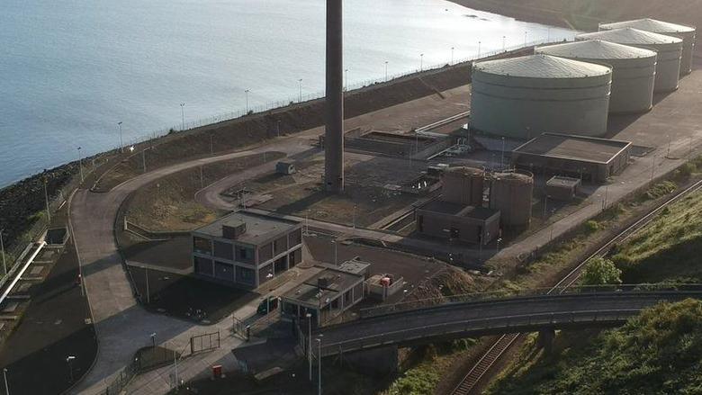 The facility has four large grey tanks.  There's a tall chimney and square shaped buildings. The sea is next to the site.