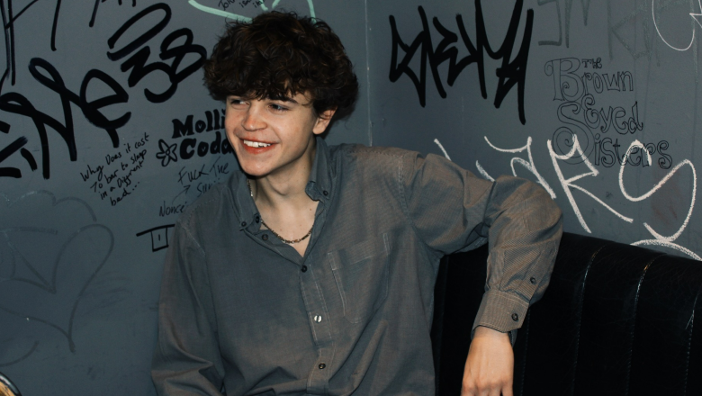 Tom A Smith, a young man with curly hair wearing a grey shirt unbuttoned to show a necklace, smiles into the distance while sitting on a black leather chair against a grey wall covered in graffiti.