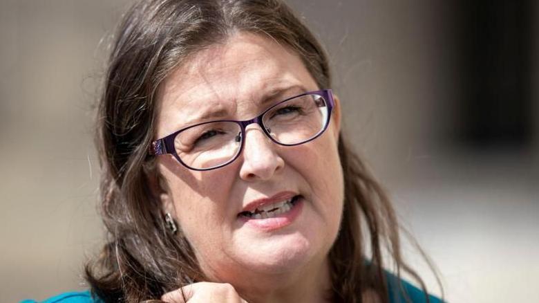 Kellie Armstrong talking to media outside Stormont. She has long brunette hair and is wearing purple framed glasses and wearing a turquoise top.