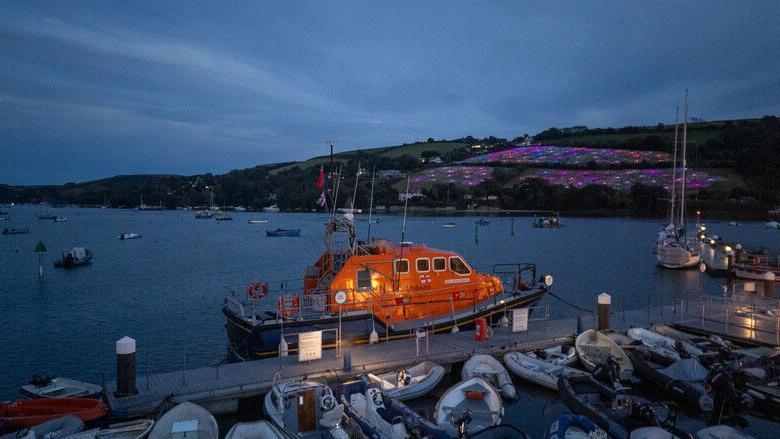 An RNLI lifeboat is tethered to the dock at dusk. In the background the Field of Light is illuminating the hillside.