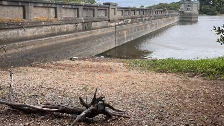 A reservoir at low level in Guernsey. A dirt bank can be seen with water further away. There is a branch in the foreground washed up on the shore. There is a large stone wall running along the left of the image.