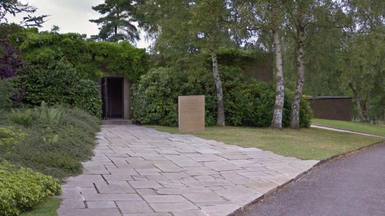 A grey, slabbed footpath that leads to a small door. Grass and trees to the right of the path, with hedges surrounding the entrance. There is a brown stone sign on the grass. 