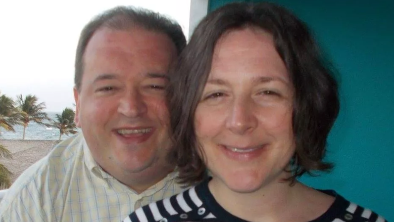 Phil Morris and his wife Dana smile at the camera. Phil has dark hair and is wearing a light yellow chequered shirt, while Dana has chin-length brown hair and is wearing a black and white striped shirt. A sandy beach and palm trees can be seen behind them. 