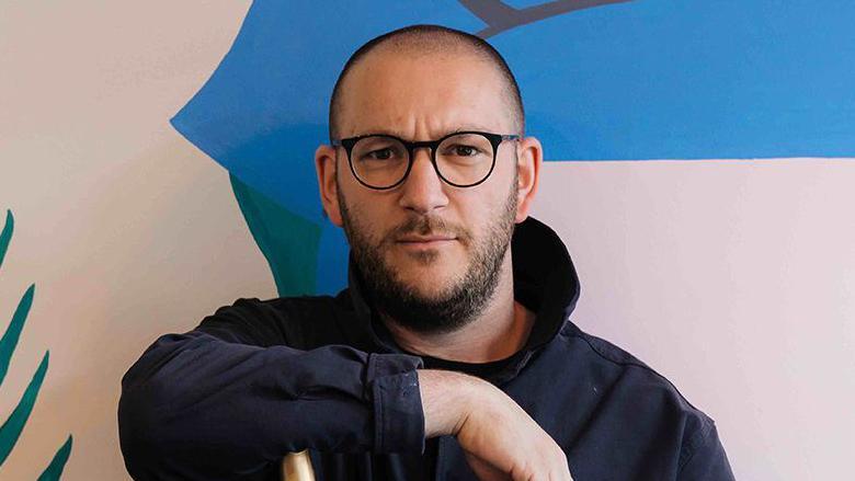 Carl Peters with short dark hair and beard and black glasses wearing a navy shirt. He is sitting in front of a feature wall which is beige blue and green.