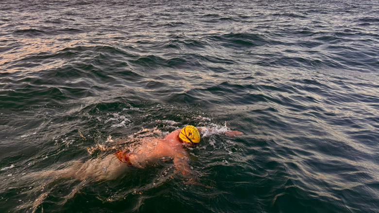 A man swimming in the English Channel at dusk. He is wearing a swim cap and goggles.