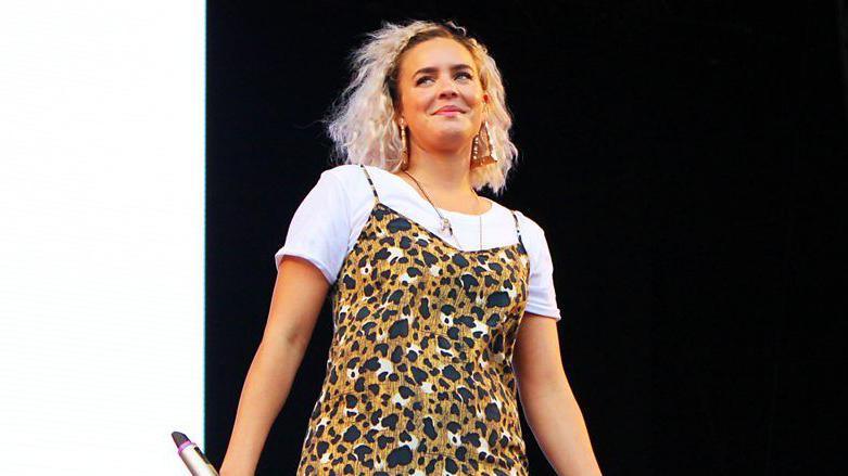 Anne Marie on stage smiling at the crowd. She has bleached blonde hair and large earrings. She is wearing a leopard-skin dress over a white top. 