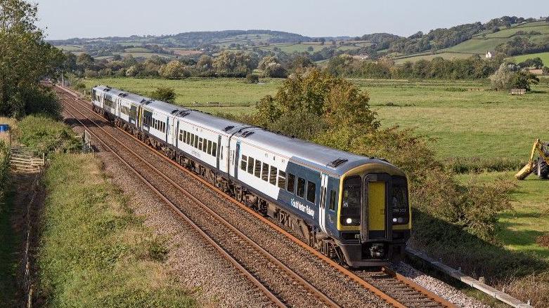 A train is travelling along a track in the countryside with trees and hills in the distance. There is grass on either side of the tracks. The sky is blue and the sun is shining.