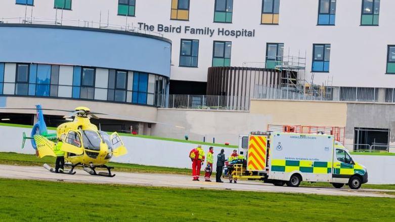 Yellow air ambulance and a person being taken into an ambulance, with a hospital in the background