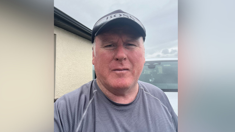 John is looking into the camera. He's wearing a grey t-shirt and aBoss cap. Behind him is a van and a building.