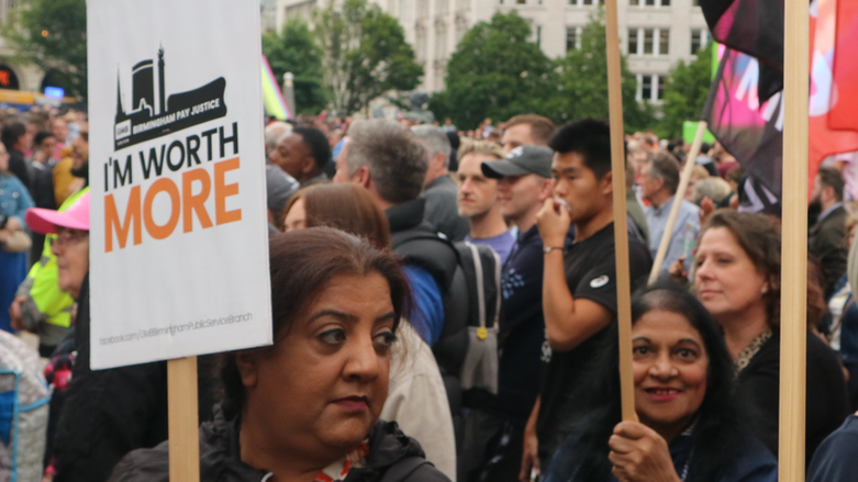 Several women holding placards with one visible saying "I'm worth more" standing next to a crowd of people.