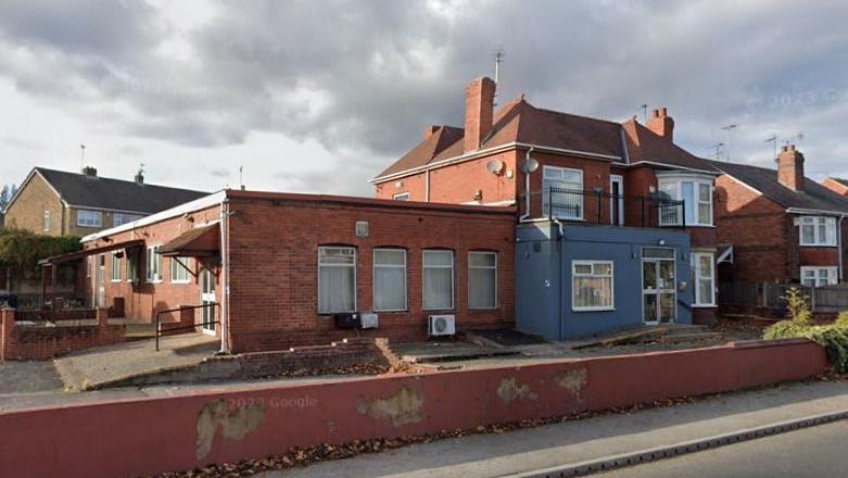 A street view image of the red brick building. The site looks empty and unused, paint is peeling off the wall in front of it. 