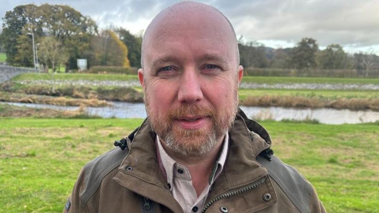 Mabon ap Gwynfor, Aelod o'r Senedd Plaid Cymru yn etholaeth Dwyfor Meirionnydd, yn edrych yn syth i'r camera tra'n sefyll yn yr awyr agored gyda chae, afon a choed yn y cefndir