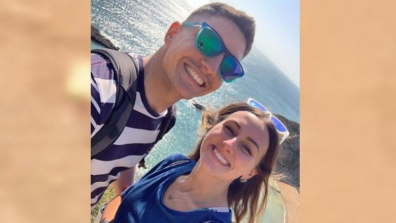 Adam Harrison and his girlfriend Chelsea smile as Adam takes a selfie. He has blue sunglasses on along with a grey backpack and blue and white striped T-shirt. Chelsea has white and blue sunglasses and a blue shirt on. They are standing in front of a beach and the sea.