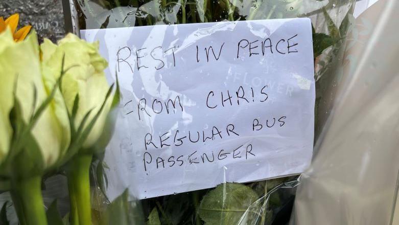 Handwritten message on yellow flowers reading: "Rest in peace, from Chris, regular bus passenger".