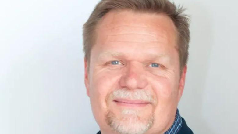 Close-up of Mr de Leeuw's face. He is smiling directly at the camera and is wearing a blue checked shirt. He has a white beard and brown hair.