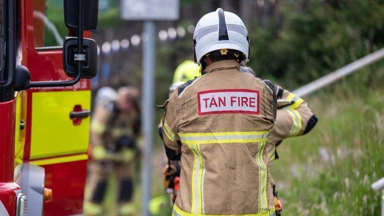 The back of a firefighter standing beside the door of a fire engine. The uniform has 'tan fire' on the back. They are surrounded by grass. 