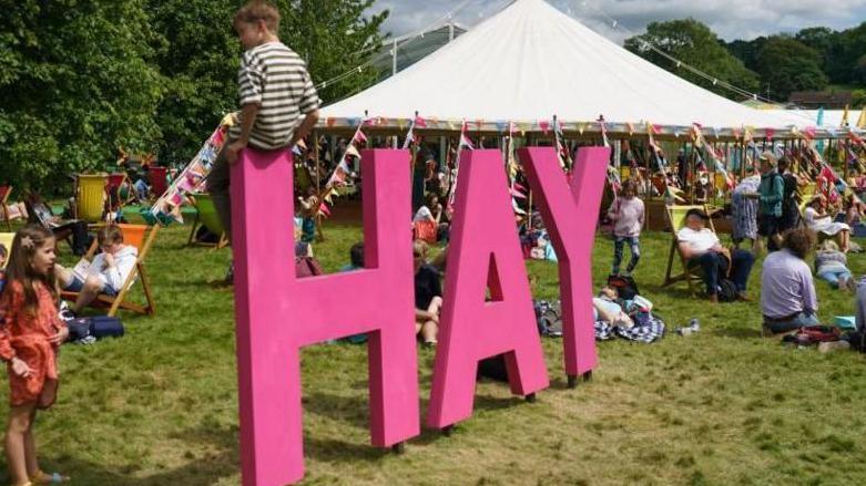 The Hay-on-Why Festival in 2022. There is large pink letter spelling out Hay while a child sits on it. The letters are surrounded by festival-goers with a large white marquee in the background.