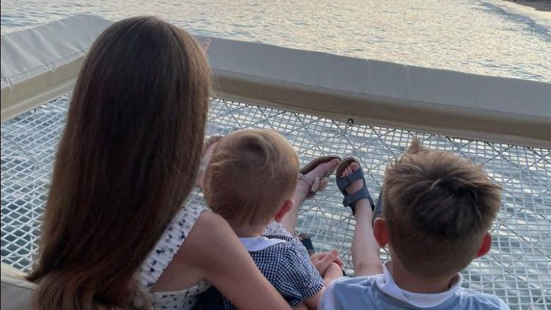 Mrs Saunders' three children sit with their backs to the camera overlooking the sunset over the see. They are sat on a large white net, directly over the water. 