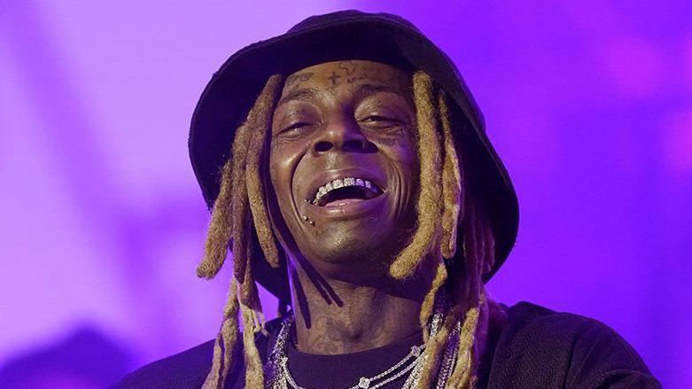 A man, smiling on stage, wearing a hat, several silver necklaces and a black top. The background is a haze of purple.