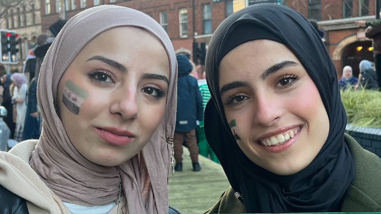 Two girls smiling, girl on left is wearing a pink headscarf, black leather jacket and cream hoody, girl on right (Renad) is wearing a black headscarf and dark green jacket, both girls have syrian flags painted on their faces, group celebrating in the background
