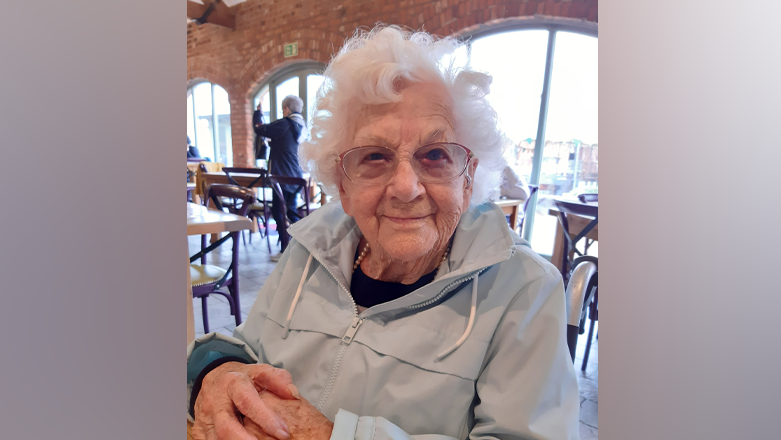 Sylvia Dalton is sitting in a restaurant and smiling at the camera. She has short white hair and pink glasses. She is also wearing a light grey coat.