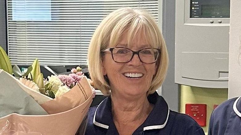 Nurse Maureen Wallis wearing a dark uniform with white piping, holding a large bouquet of flowers wrapped in paper. Behind the person is an indoor setting with a window featuring horizontal blinds, a wall-mounted control panel, and a red emergency button.