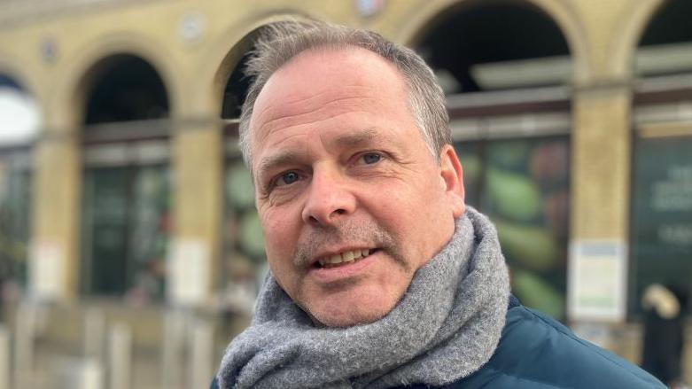 A man standing outside Cambridge station. He is smiling at the camera and has cropped grey hair. He is wearing a grey scarf and blue jacket. 