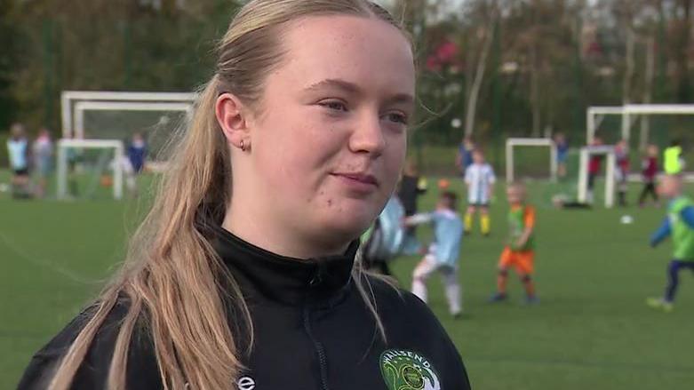 Daisy with long blonde hair talks to the camera. Behind her is a blurred background of children playing football in different colours of kit.