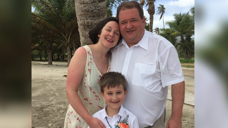 Phil, Dana and their young son Orson smile at the camera. Phil and Orson are both wearing white shirts, while Dana is wearing a yellow dress with red flowers on. There are palm trees in the background. 