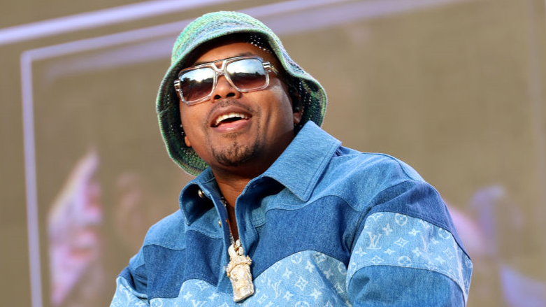 US rapper Nas wearing sunglasses and a hat smiles on stage in a blue shirt 