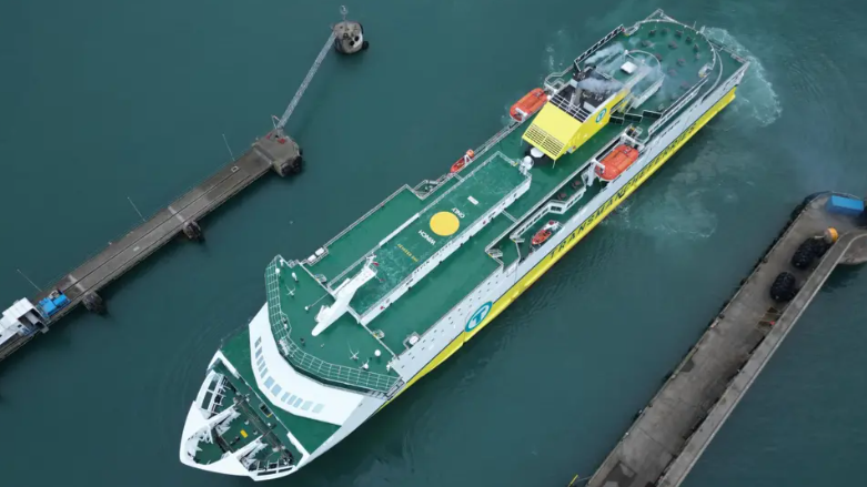 An aerial image of a DFDS ferry on the water. It has a largely green floor surface, and white and yellow sides. 