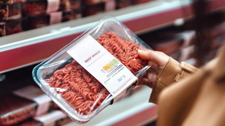 A shopper holds a packet of beef mince in her hand in front of a supermarket shelf.
