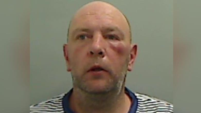 A mugshot of Paul Sizer who is looking to the side of the camera. He is bald, with brown and grey stubble and is wearing a blue and white stripped t-shirt.