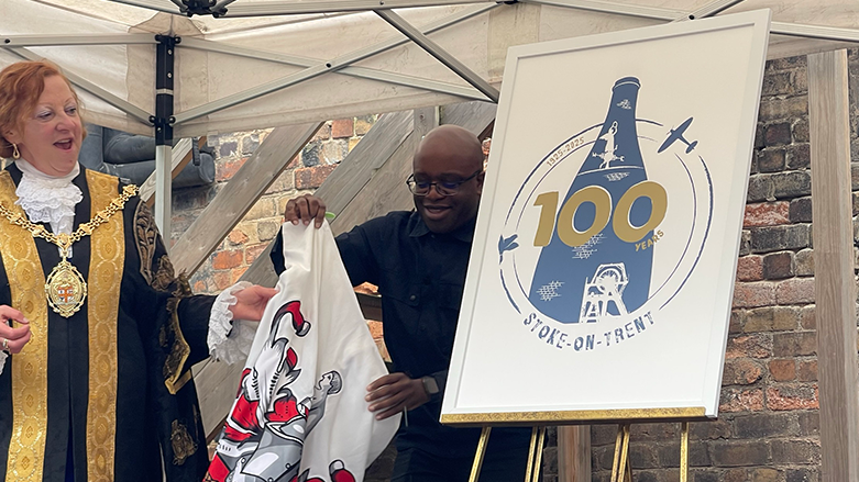 Stoke-on-Trent Lord Mayor Lyn Sharpe, in black and gold mayoral robes, is seen unveiling a logo which features a traditional bottle oven and the number 100. Next to her is Reverend Geoff Eze holding a white sheet with the city's crest on it.