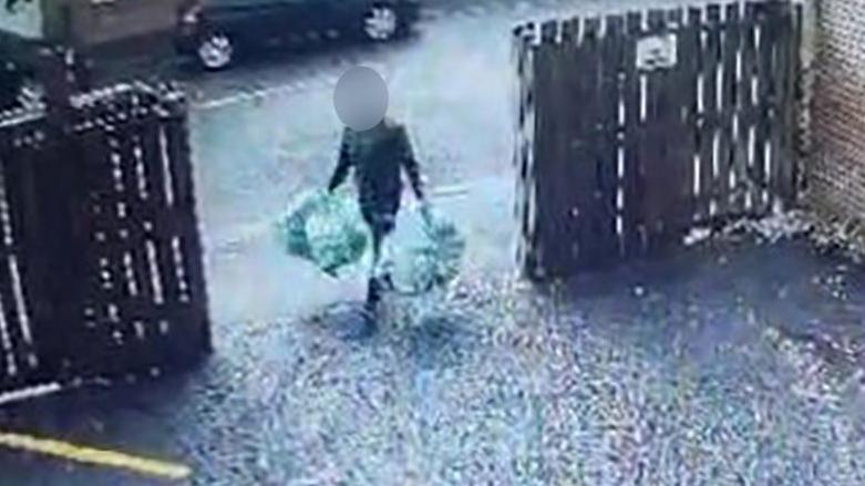 A man carrying four bags of rubbish, two in each hand. He is walking through an open gate into a driveway, with wooden fencing either side. His face is obscured. 