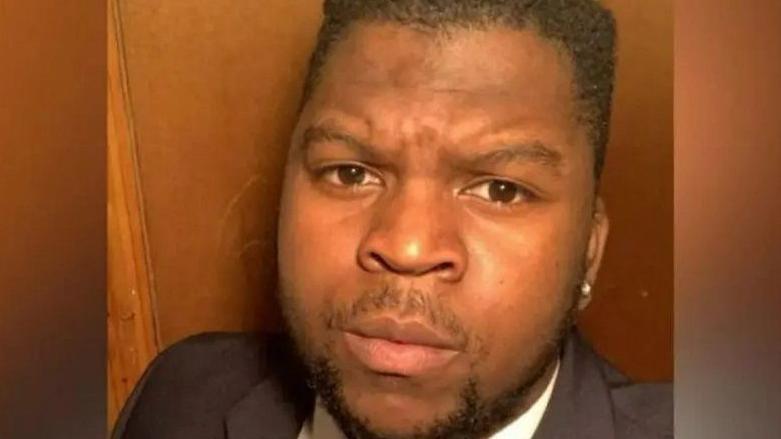 Saymore T Kwashira looking at the camera with a serious expression. He has a black beard and black hair and has a white shirt and black jacket on, stood in front of a wooden background.