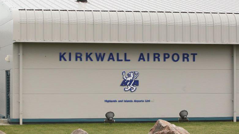 A general view of Kirkwall Airport. The terminal building is grey, with the name and a lion motif in blue and white on the side. There is a patch of green grass in front.