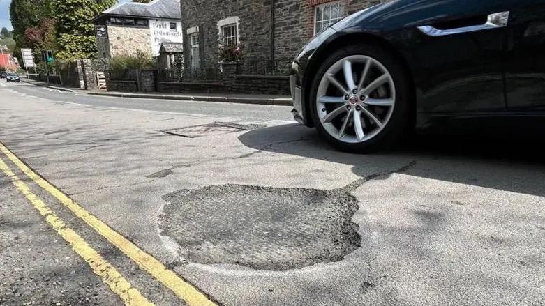 A pothole on Dolvin Road in Tavistock with a passing navy blue car.