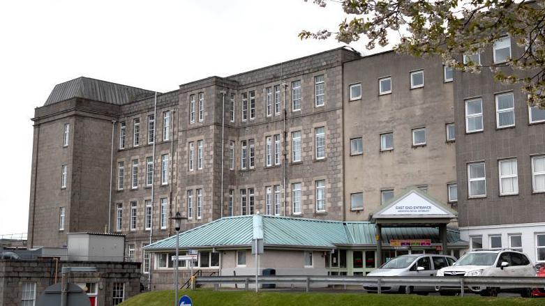 Grey hospital building with cars parked outside, a top of a grass verge.