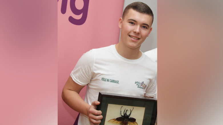 A young Liam Óg Ó hAnnaidh, known by his stage name Mo Chara, smiles at the camera. He is wearing a white tshirt with the Glór na Móna logo on one side and Féile Na Carraige on the other. He is holding a framed bug and is standing in front of a pink backdrop.