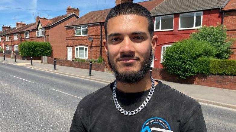 Young man with a beard and dark hair, eyes and skin, wearing a black t-shirt and silver chain, standing on a street in Maltby, Rotherham with a pavement and red brick houses and a hedge behind.